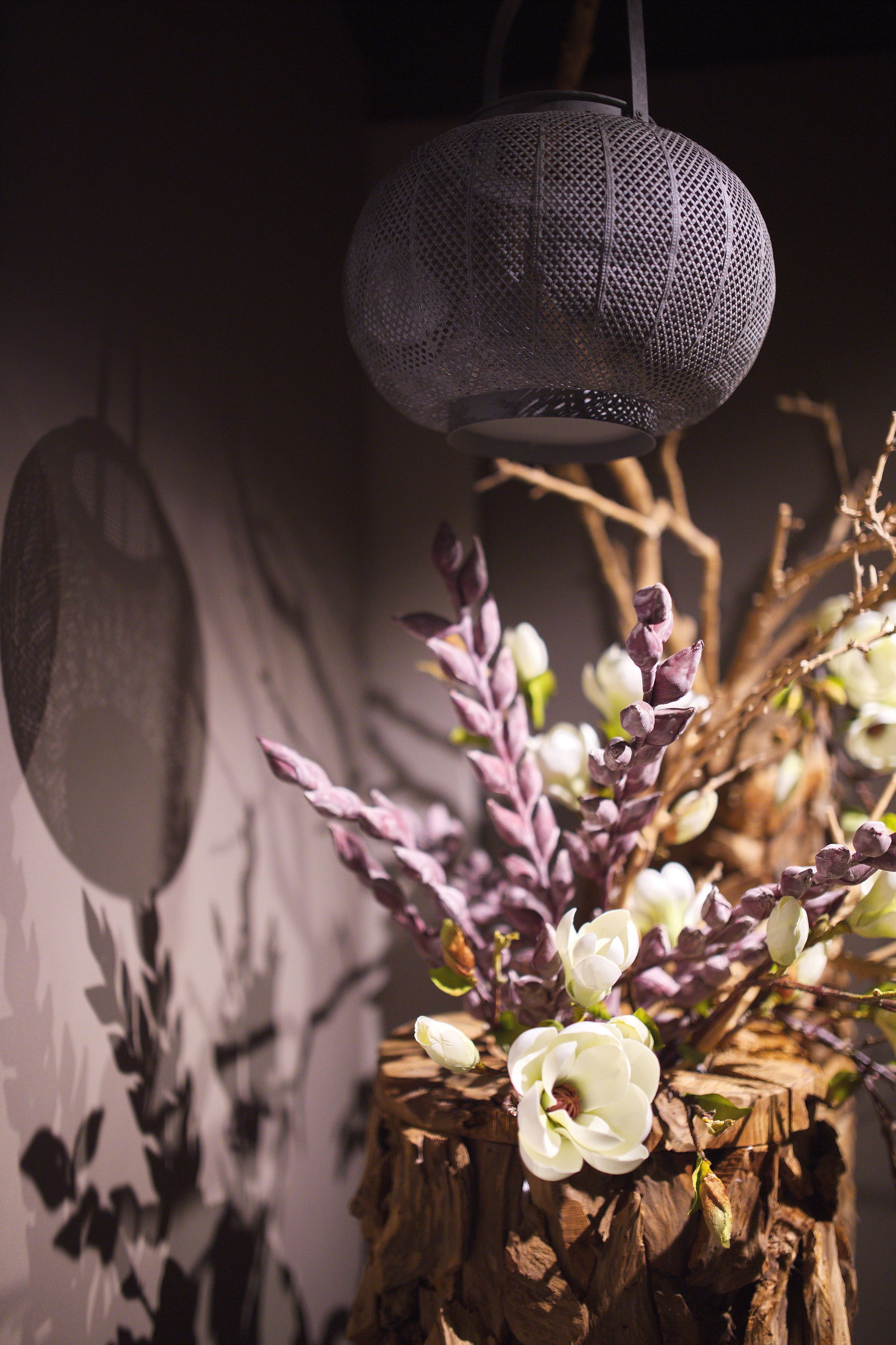 Une lanterne suspendue à la surface texturée, dotée d’un socle en bois brut et agrémentée d’une composition florale aux fleurs blanches et violettes.