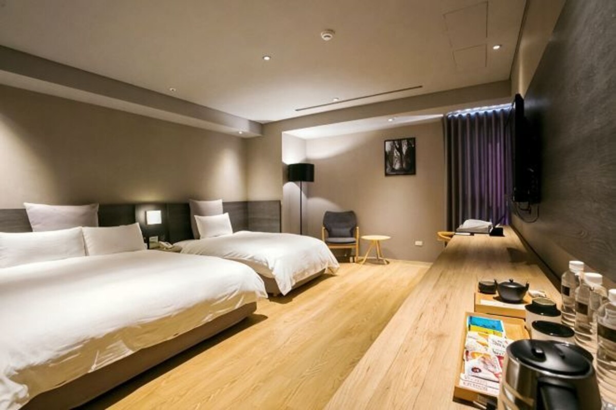 Quarto de hotel com duas camas, uma escrivaninha de madeira com comodidades, uma cadeira e uma televisão na parede.