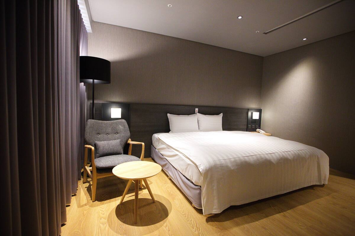A hotel room with a large bed, a small round table, a chair, a lamp, and a nightstand with a phone.