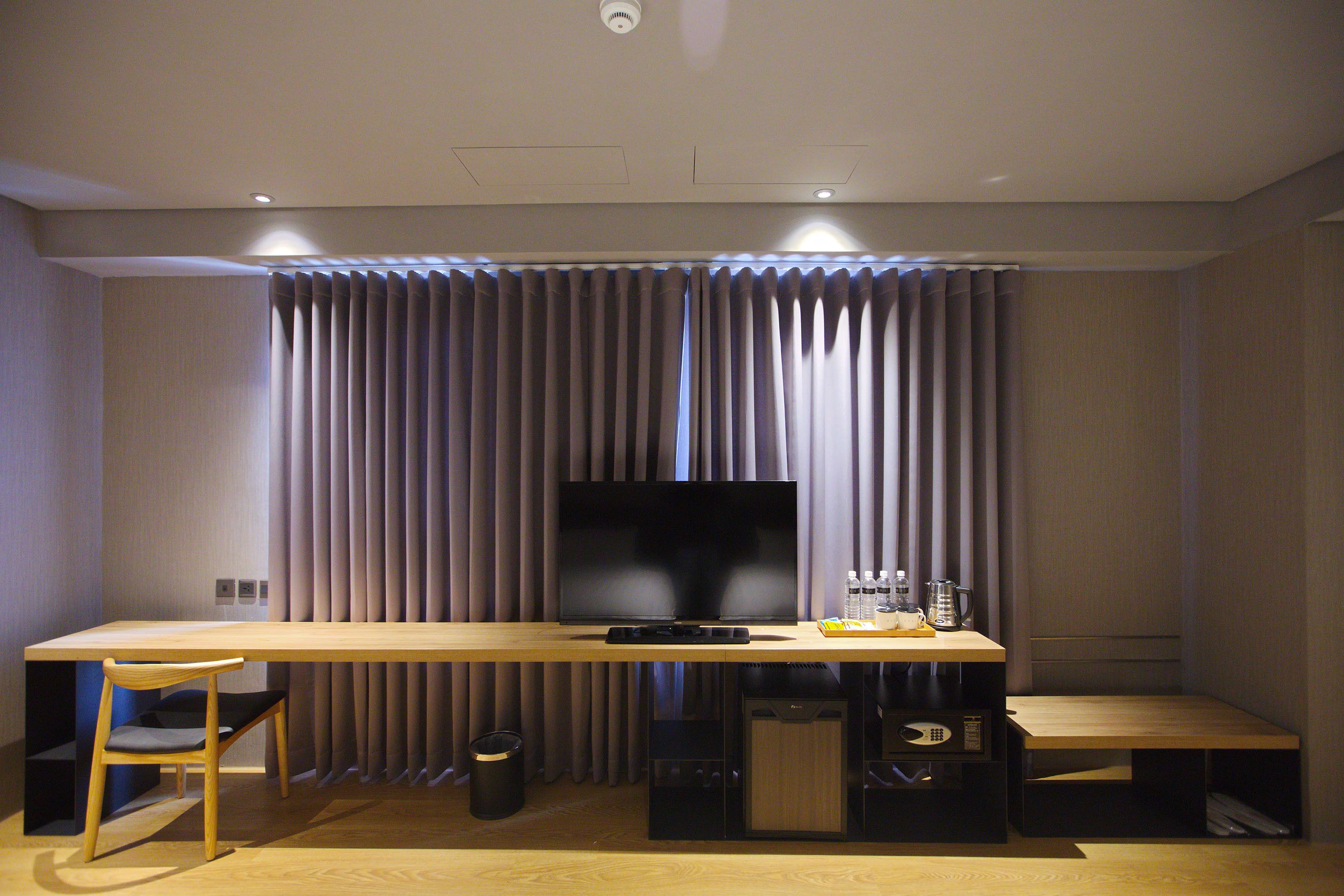 Een moderne hotelkamer met een houten bureau, een flatscreen televisie, een stoel, een bankje en een gordijnenachtergrond.