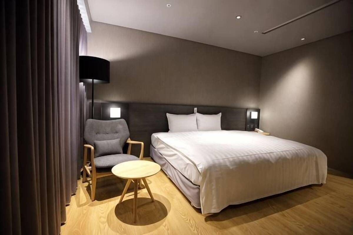 A hotel room with a bed, a chair, a small table, and a lamp.