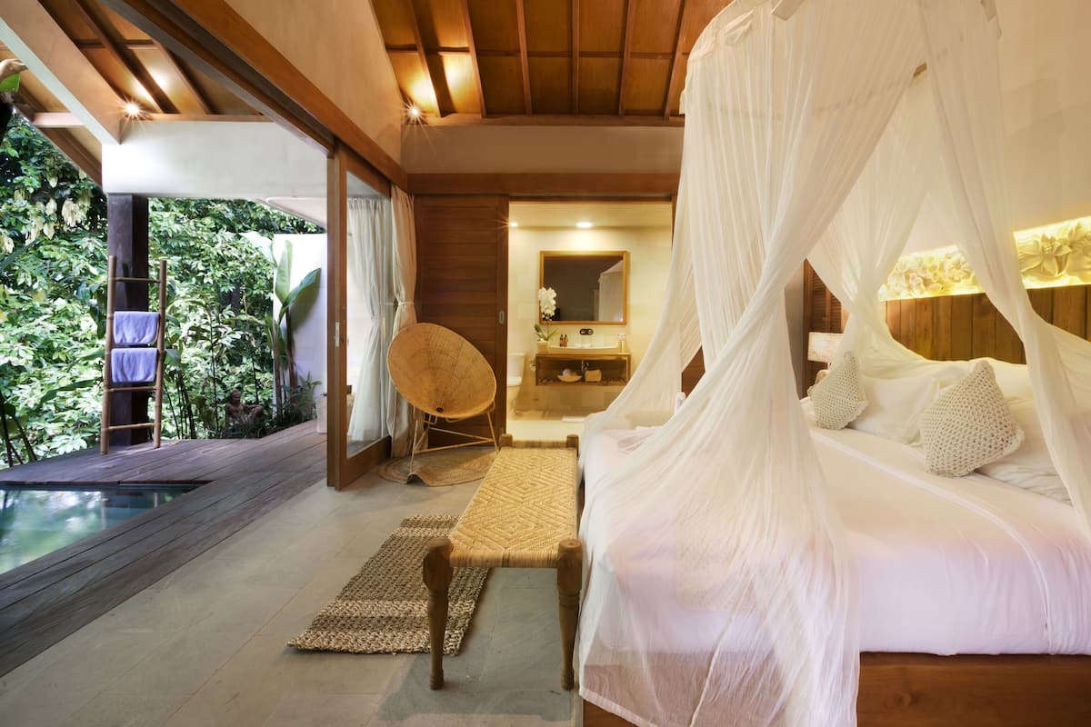 A bedroom with a canopy bed, a wooden floor, a view of a pool, and a bathroom with a mirror and sink.