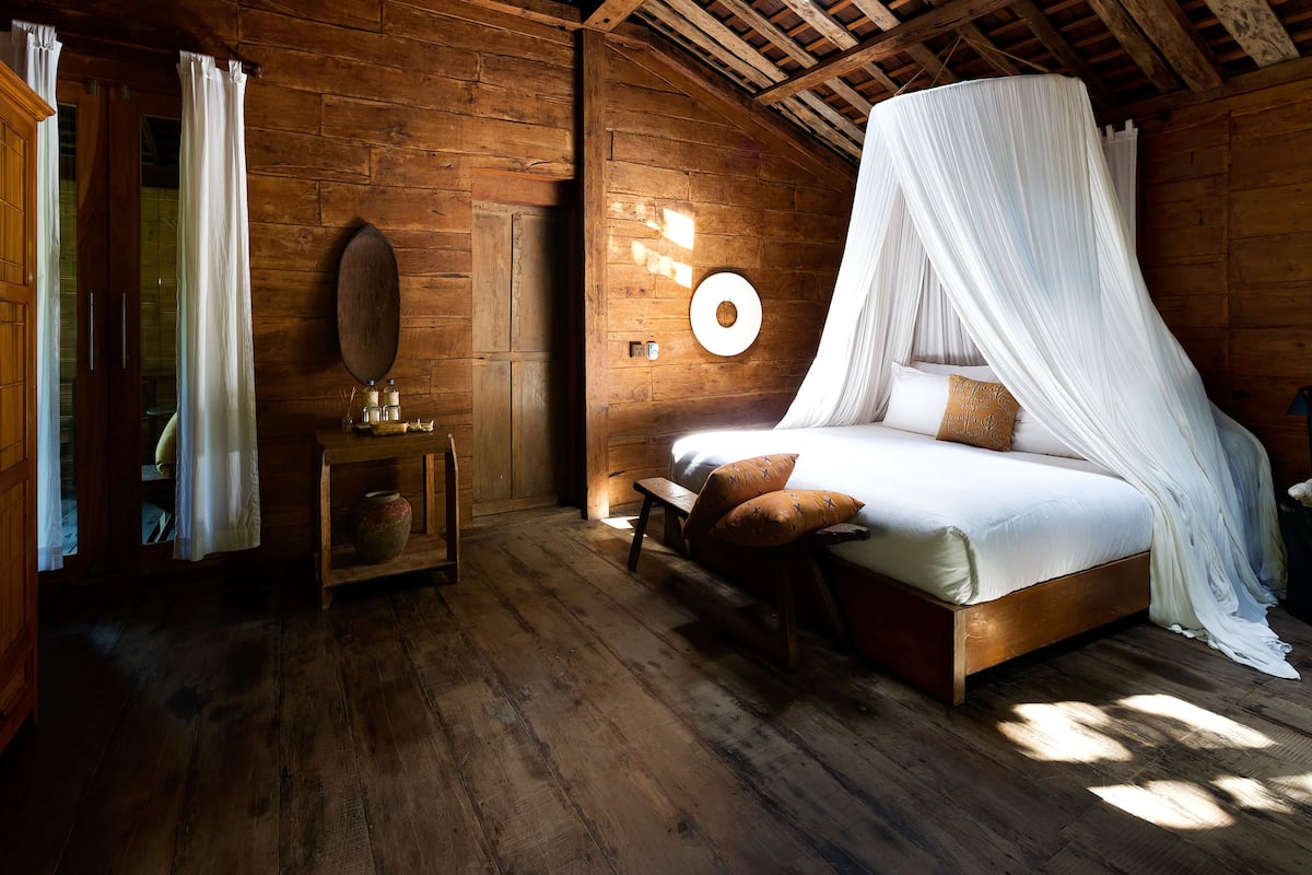 A wooden cabin bedroom with a bed, a nightstand, a mirror, and a window with curtains.