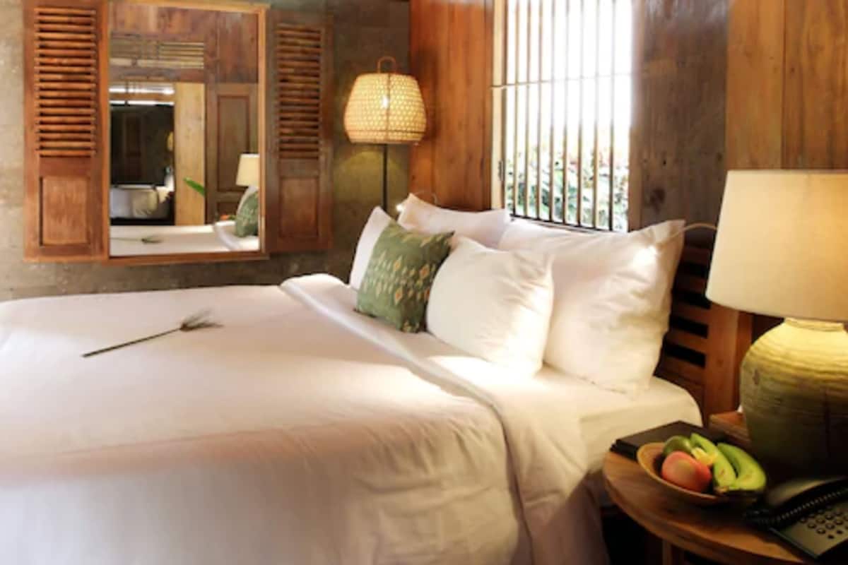 A neatly made bed with white linens and a single decorative item, a wooden headboard with a mirror, a wicker lamp, and a small table with a fruit bowl and a telephone.