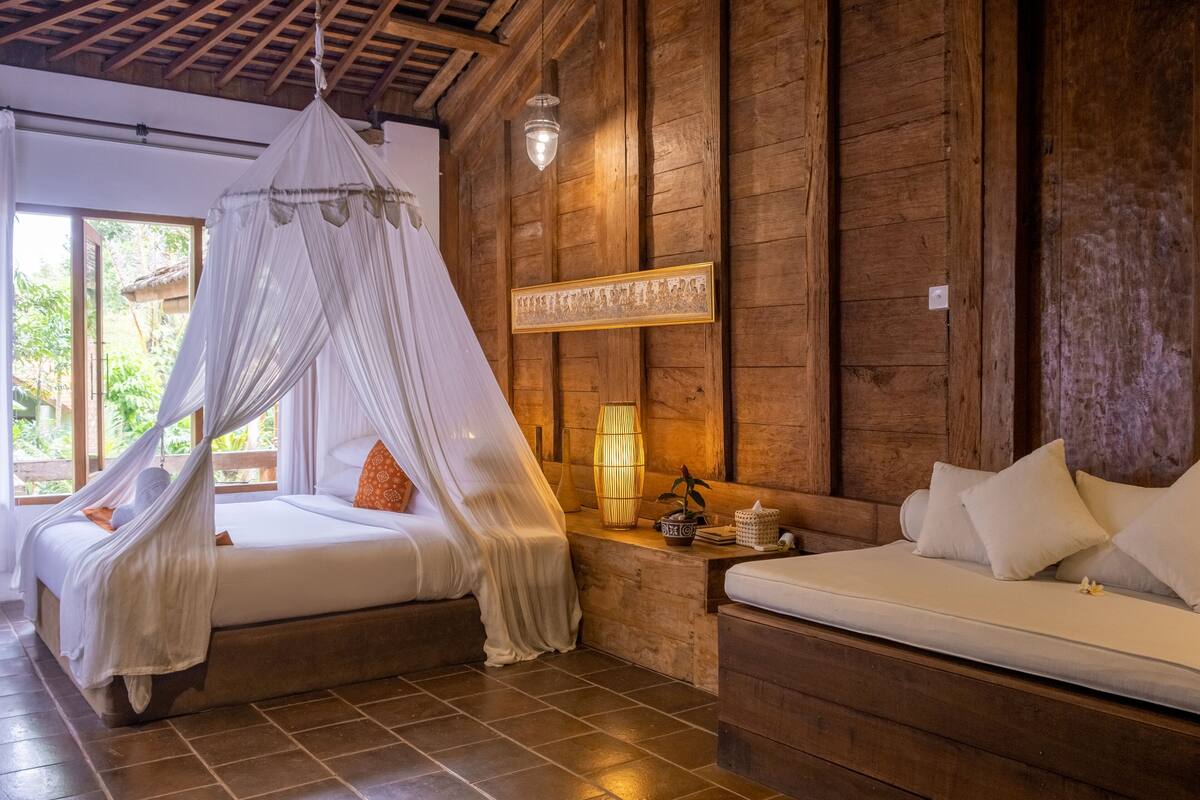 A room with a bed under a canopy, a sofa, wooden walls, a bedside lamp, and a window with a view of greenery.