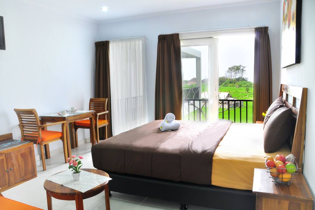 A hotel room with a bed, a desk with a chair, a small table with a plant, and a balcony with a view of greenery.