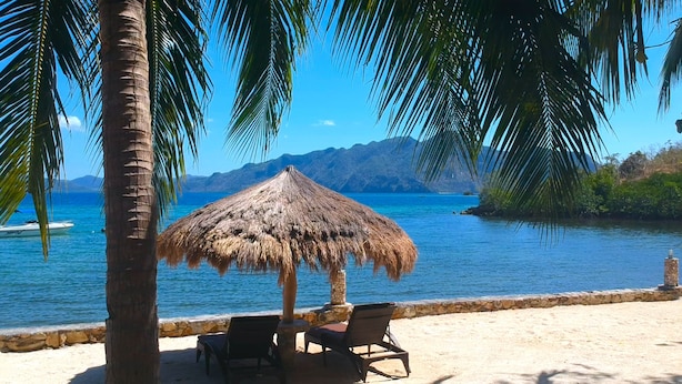 A beach with a thatched umbrella, two lounge chairs, and a view of the sea and mountains.