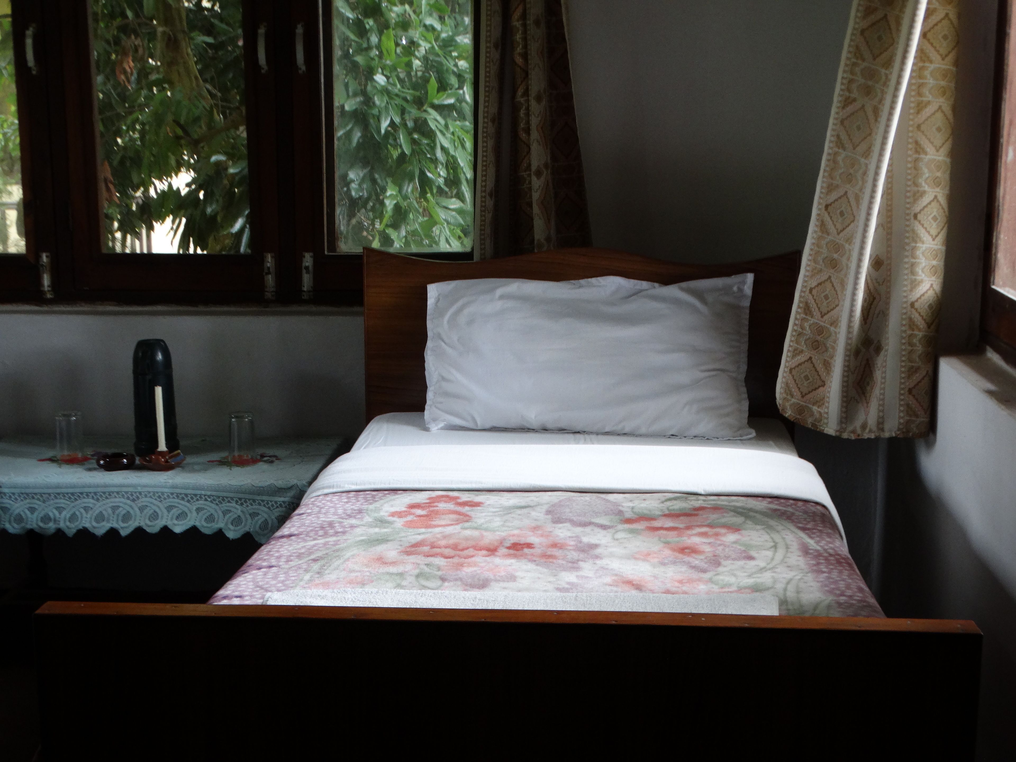 A bed with a floral bedspread, a white pillow, and a wooden headboard. There is a small table with a lace tablecloth and a black object on it.