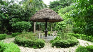 A thatched-roof gazebo with a table and chairs in a lush garden setting. - Nepal Wildlife Resort (Thakudwara)