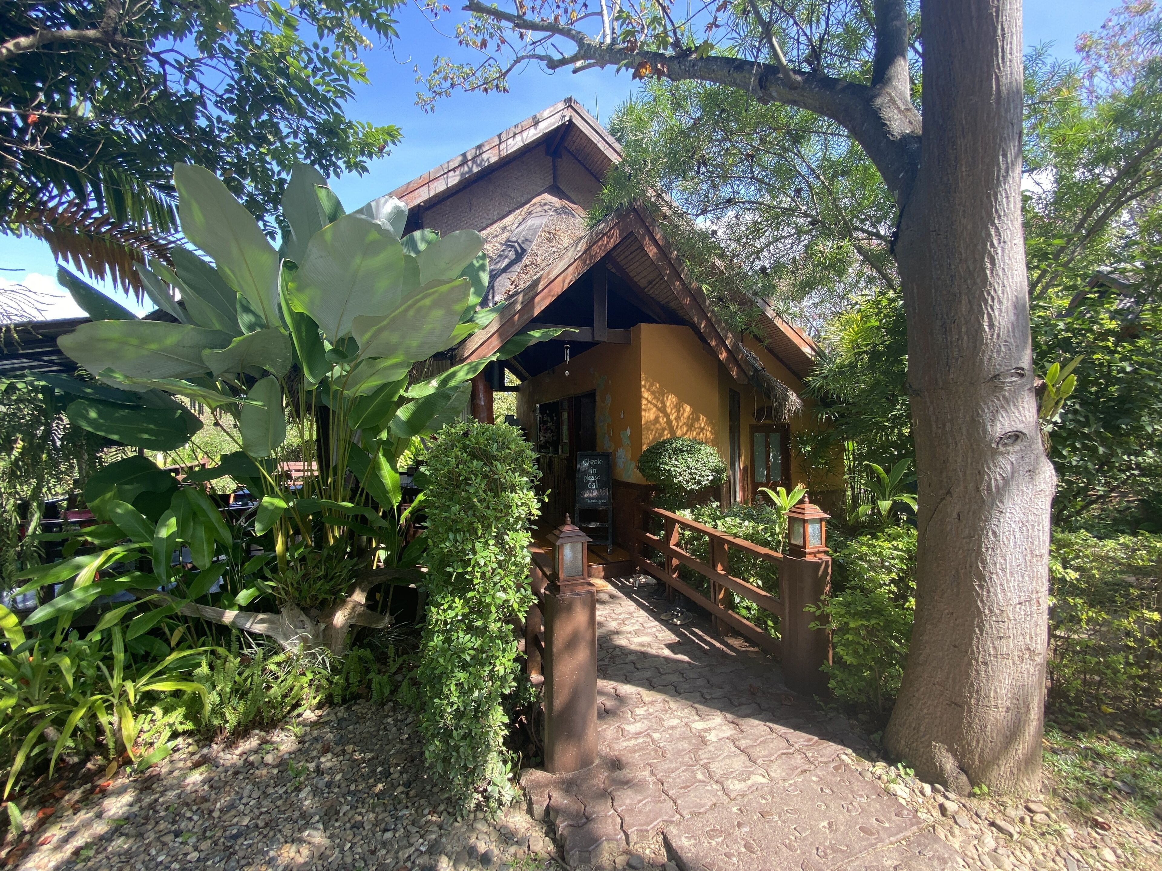 A rustic building with a thatched roof, surrounded by lush greenery and a cobblestone path leading to the entrance.