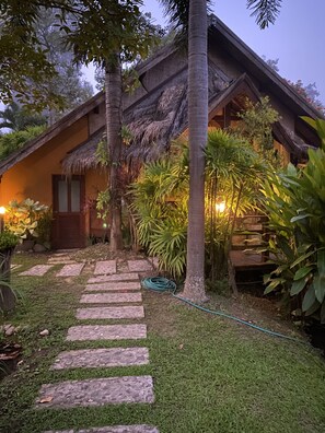 A tropical-style building with a thatched roof, surrounded by lush greenery and a stone pathway leading to the entrance.