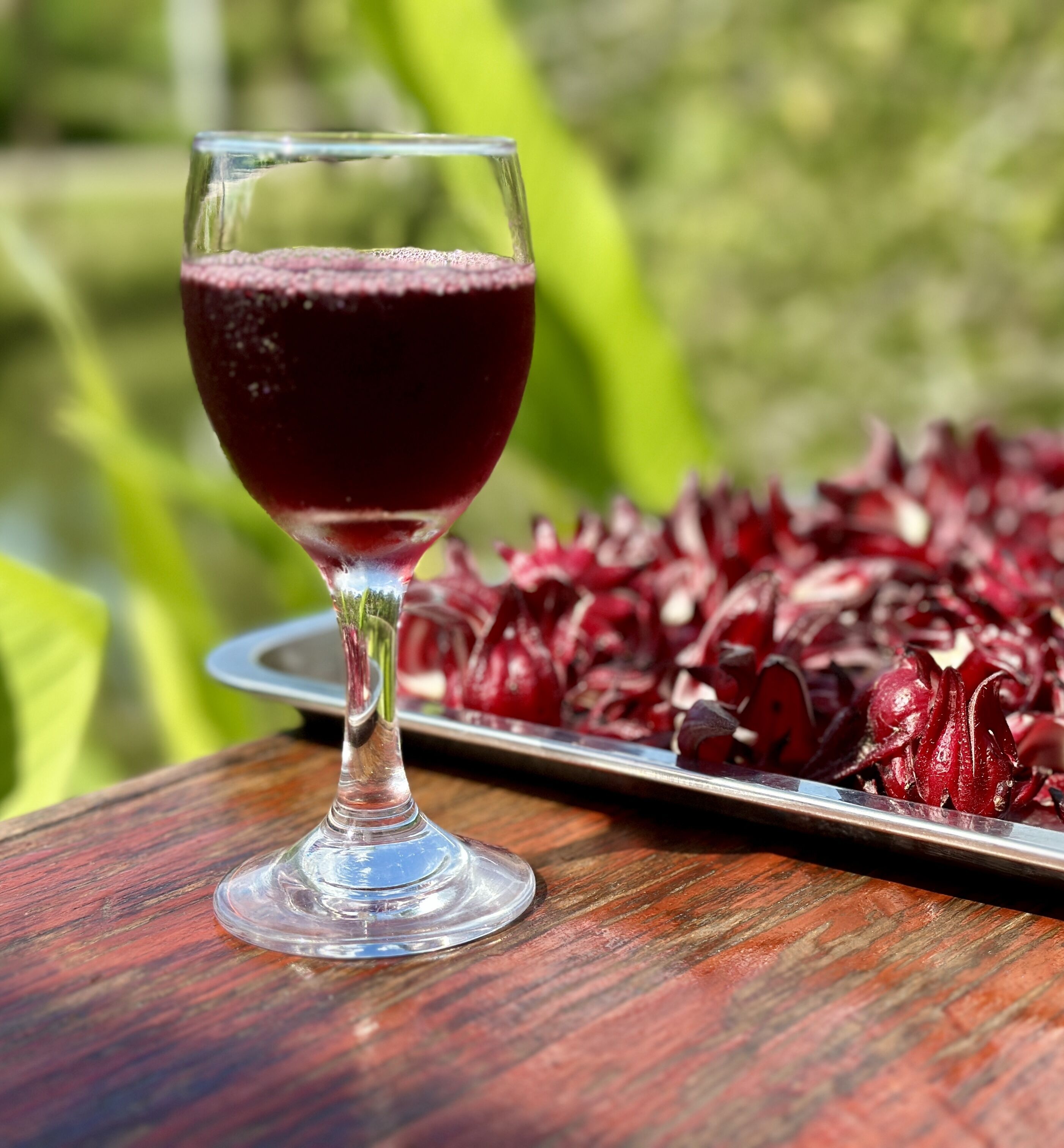 A glass of red beverage on a wooden table with a tray of red fruits in the background.