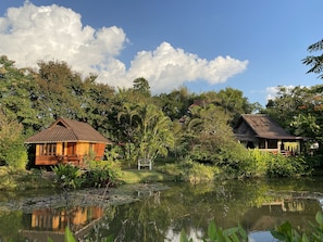 A rustic wooden house with a thatched roof, surrounded by lush greenery and a calm pond.