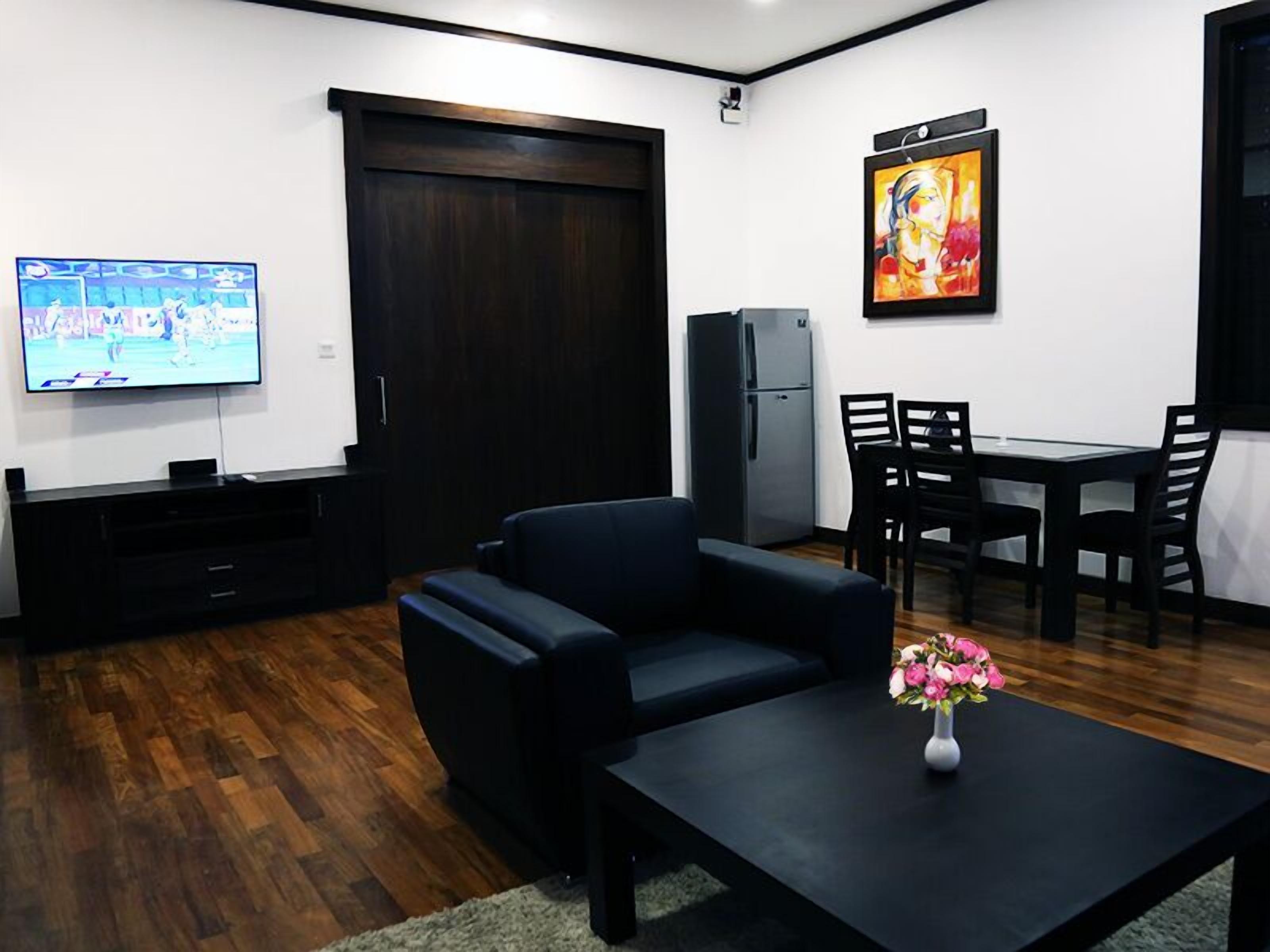A modern living room with a dark wooden floor, a black sofa, a glass coffee table with a vase of flowers, a flat-screen TV, a dark wooden door, a refrigerator, and a dining area with a table and chairs.