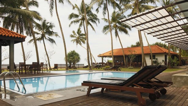 A swimming pool with lounge chairs and a covered area, surrounded by palm trees and a building with a tiled roof. - Blue Beach (Wadduwa)