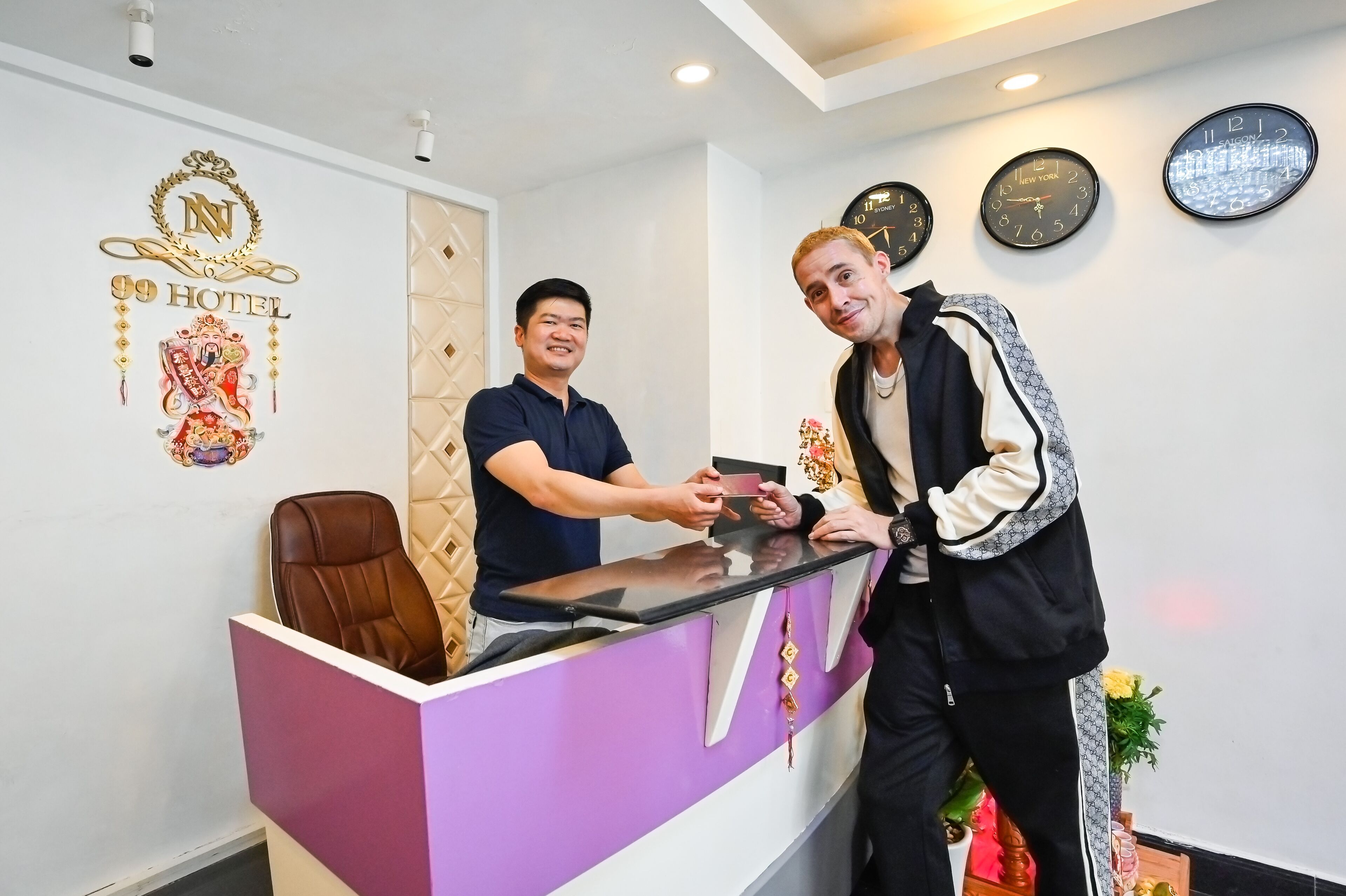 Un comptoir d’accueil d’hôtel avec un comptoir violet, une chaise marron et une horloge affichant l’inscription « New York » sur le mur.