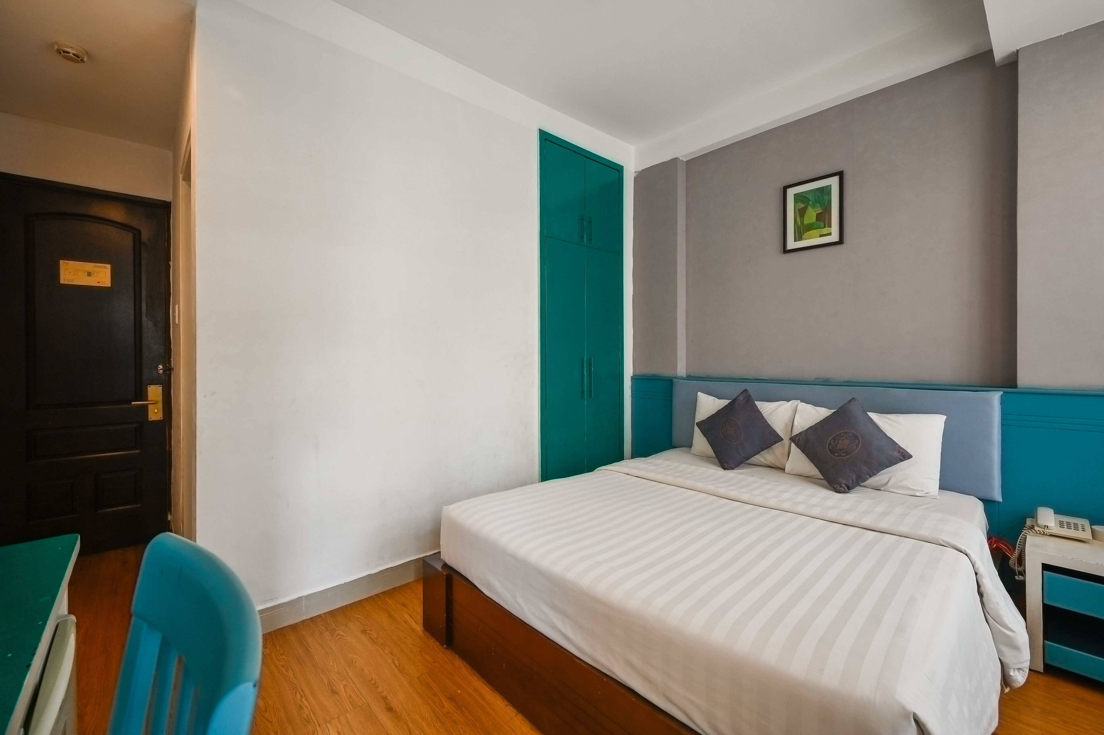 A hotel room with a bed, a desk, a chair, a phone, and a door.