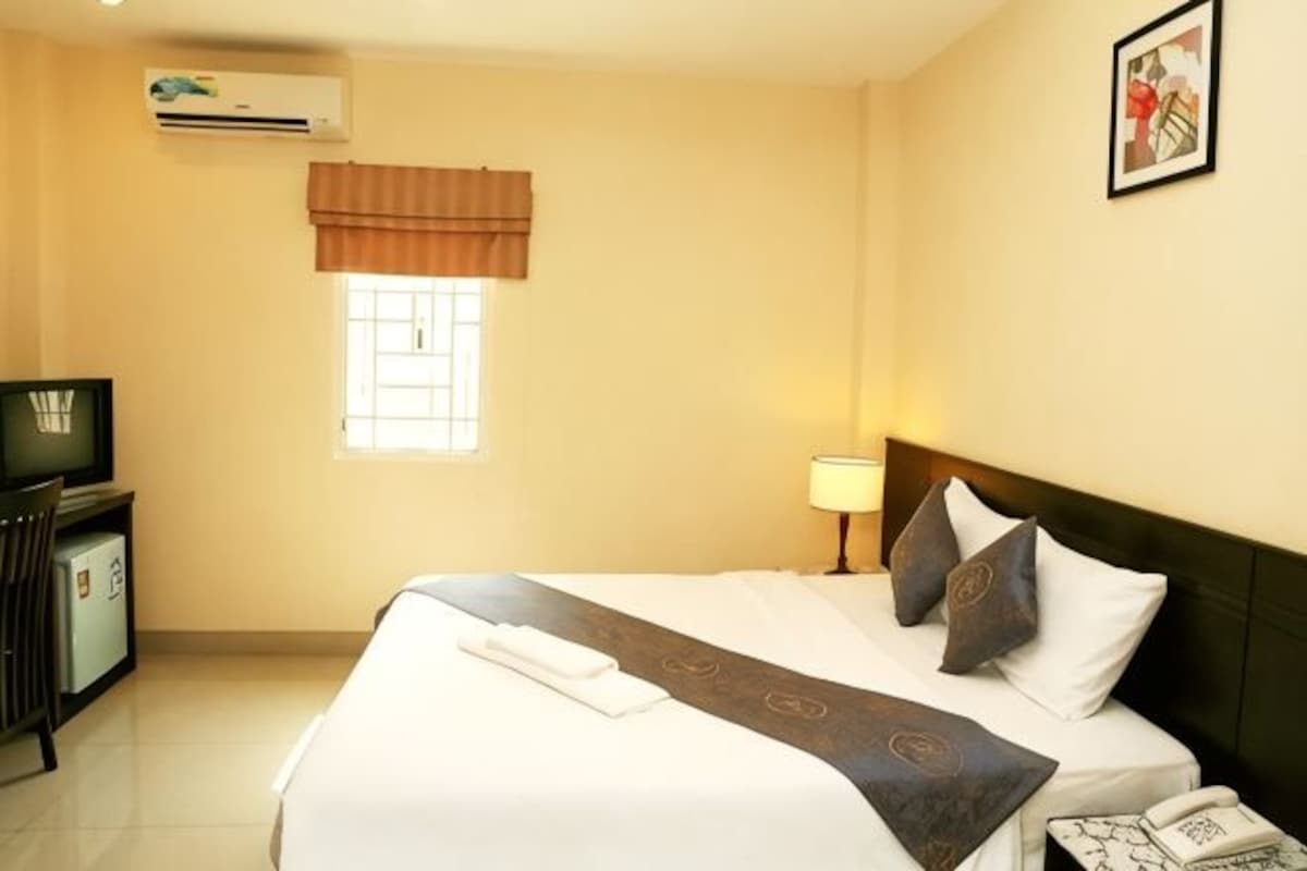 Una habitación de hotel con una cama, un escritorio con computadora, una lámpara y un ventanal con cortinas.