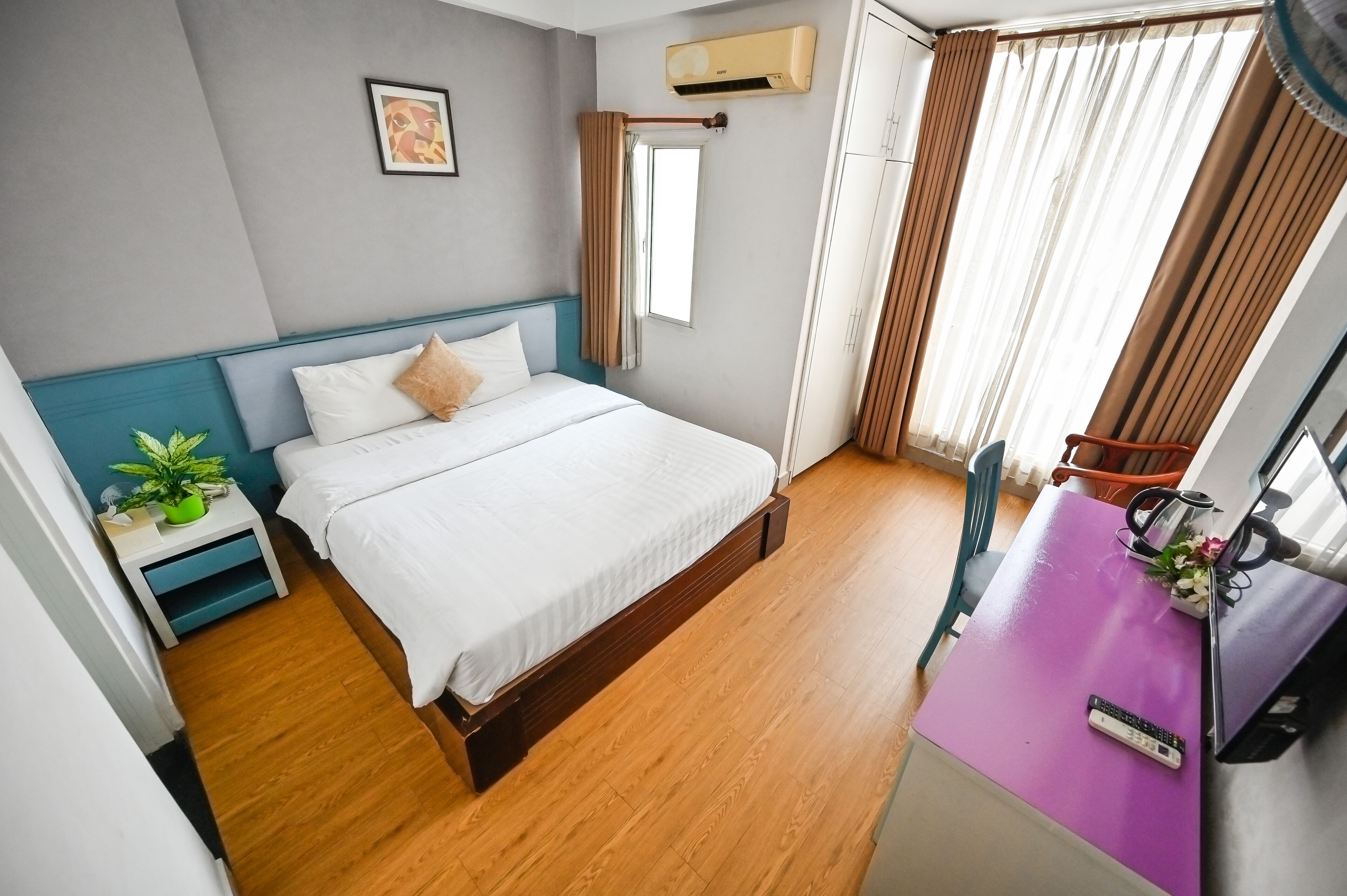 Habitación de hotel con cama, escritorio con computadora, ventana con cortinas, aire acondicionado y una planta pequeña en la mesita de noche.