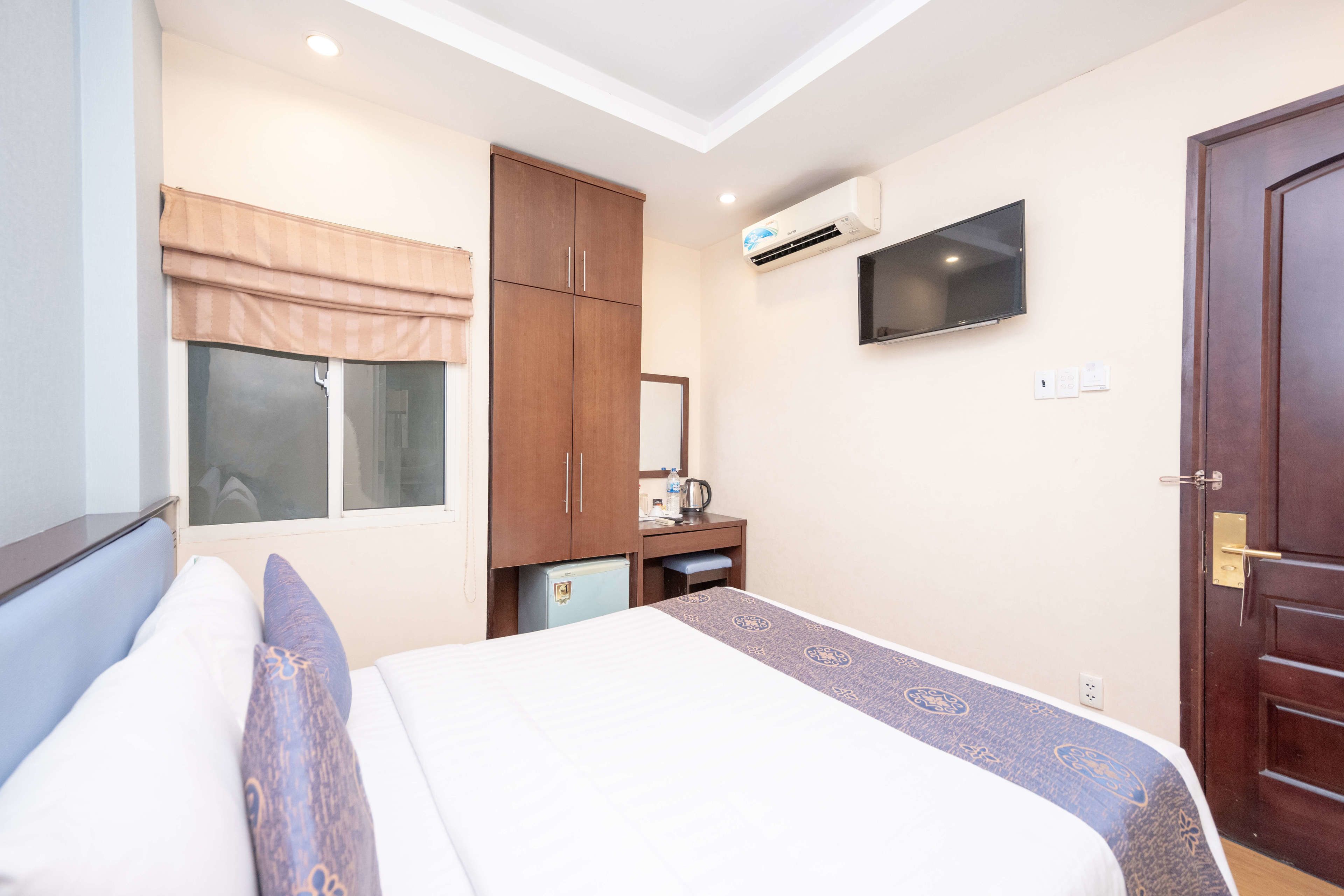 A hotel room with a bed, a wardrobe, a desk with a chair, a television, and an air conditioning unit.