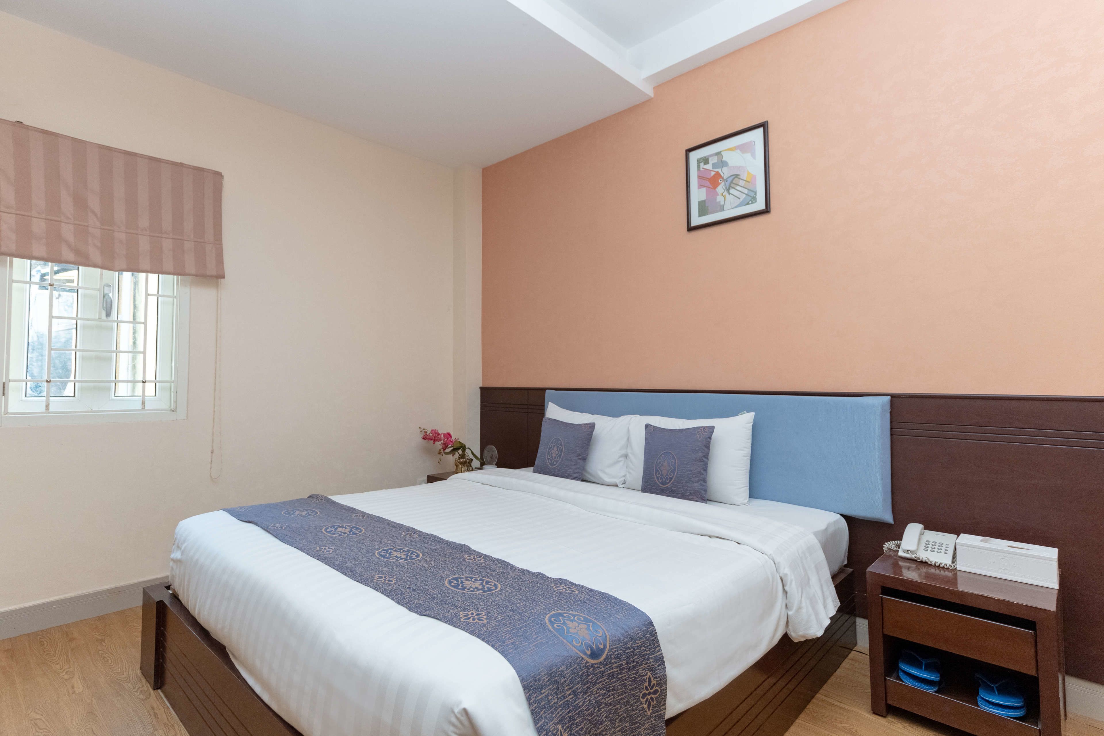 A neatly made bed with a blue and white bedspread, a wooden nightstand with a phone and a lamp, and a window with a beige curtain.