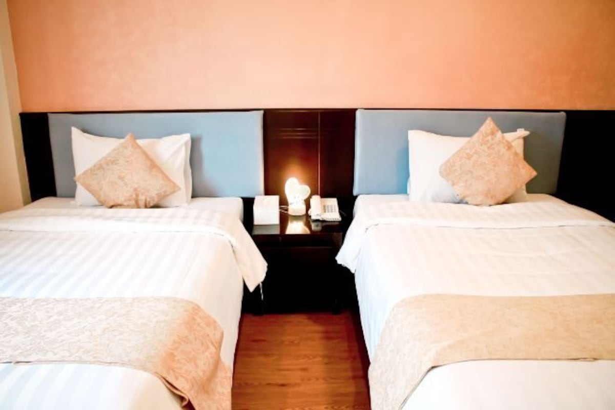 Habitación de hotel con dos camas, cada una con cabecera, mesita de noche con lámpara y teléfono fijo en la pared.