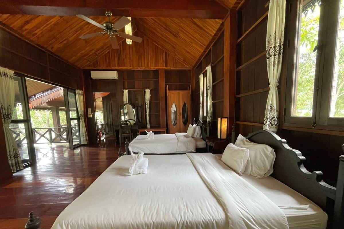 A hotel room with two beds, wooden flooring, a ceiling fan, and large windows with curtains.