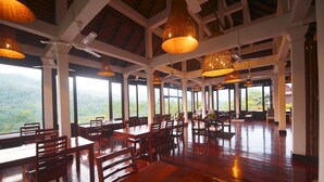 A spacious dining area with wooden tables and chairs, large windows offering a view of the mountains, and hanging wicker lamps. - Nam Ou Riverside Hotel & Resort (Pak Ou)