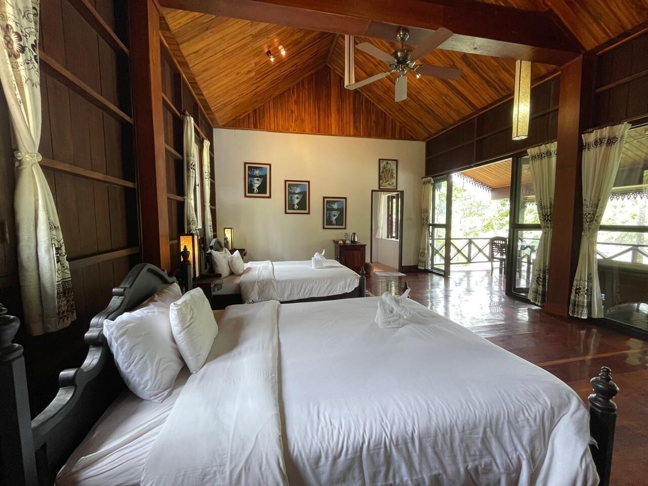 A large, well-lit room with two beds, wooden ceiling beams, and large windows offering a view of the outdoors.