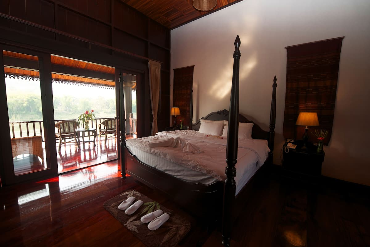 A four-poster bed with a wooden headboard, a nightstand with a lamp, and a view of a balcony with wicker furniture.