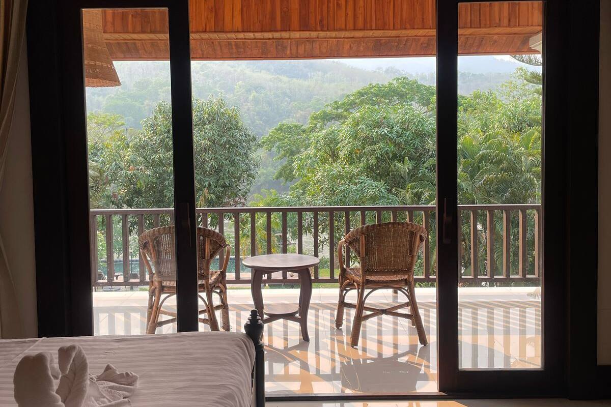 A hotel room with a bed, a small table, and chairs. The room has a balcony with a view of greenery and a wooden ceiling.