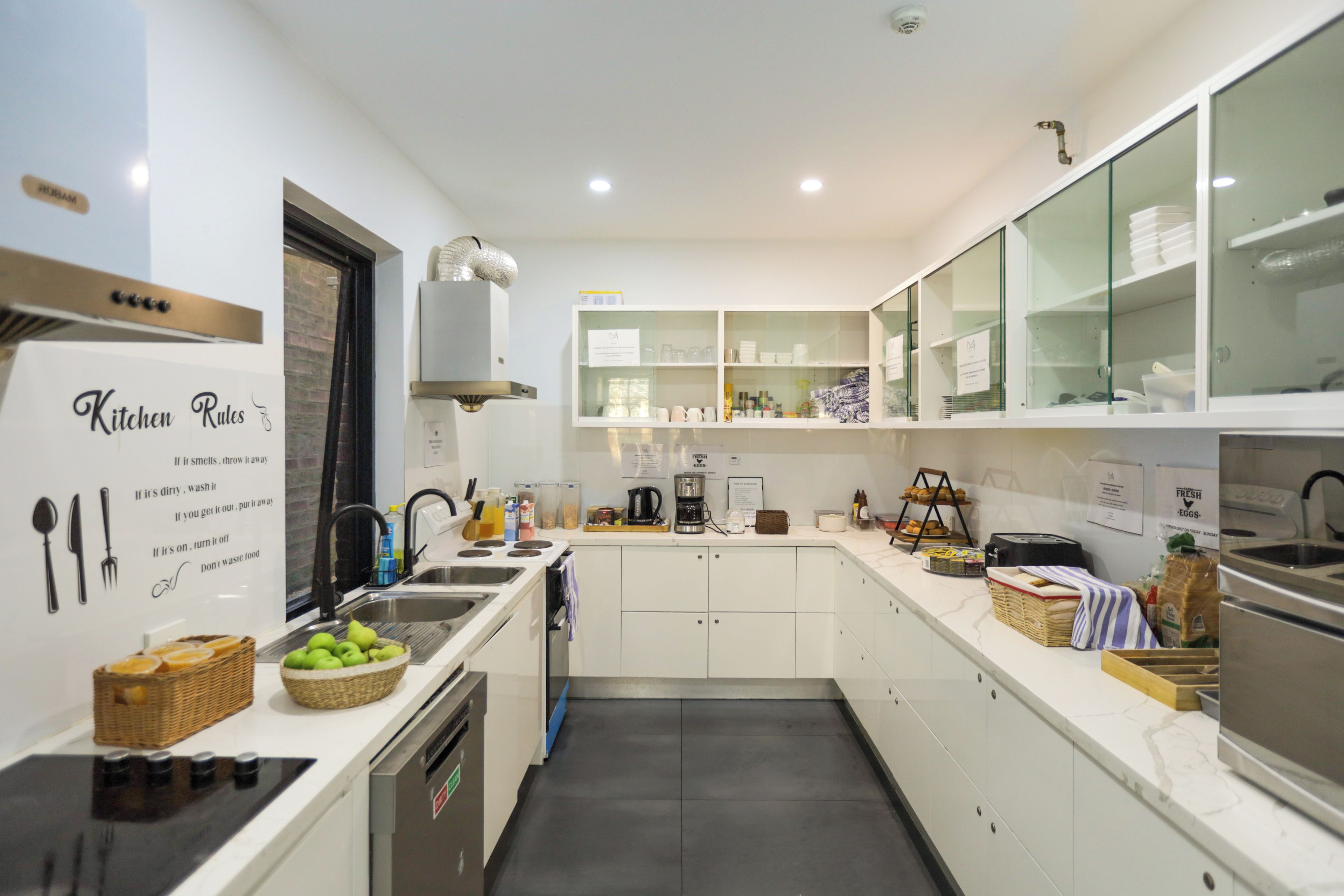 Una cocina moderna con armarios blancos, horno empotrado, pileta y extractor. Hay cestas con fruta y una cafetera en la encimera.