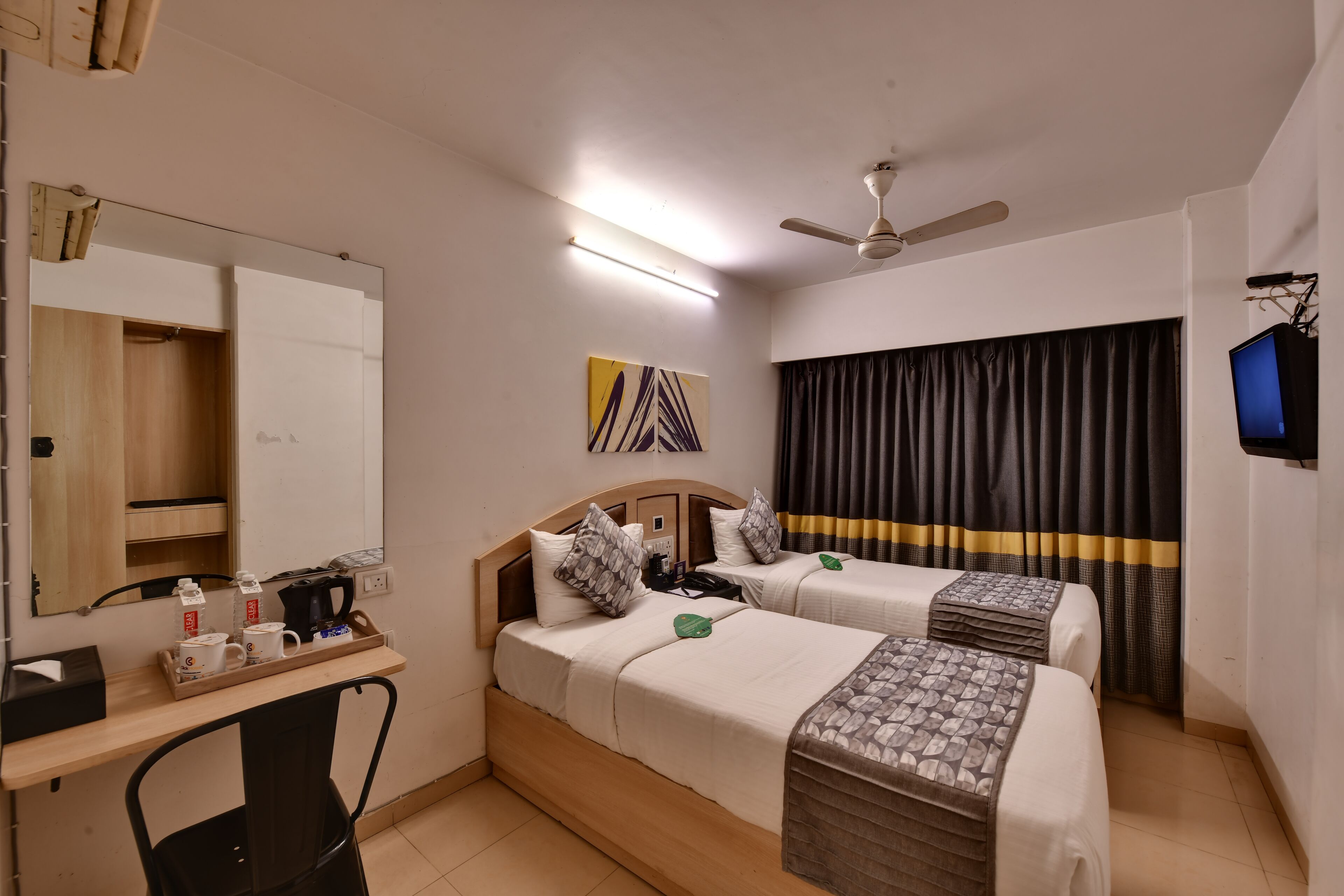 A hotel room with two beds, a desk with a chair, a ceiling fan, a flat-screen TV, and a window with curtains.