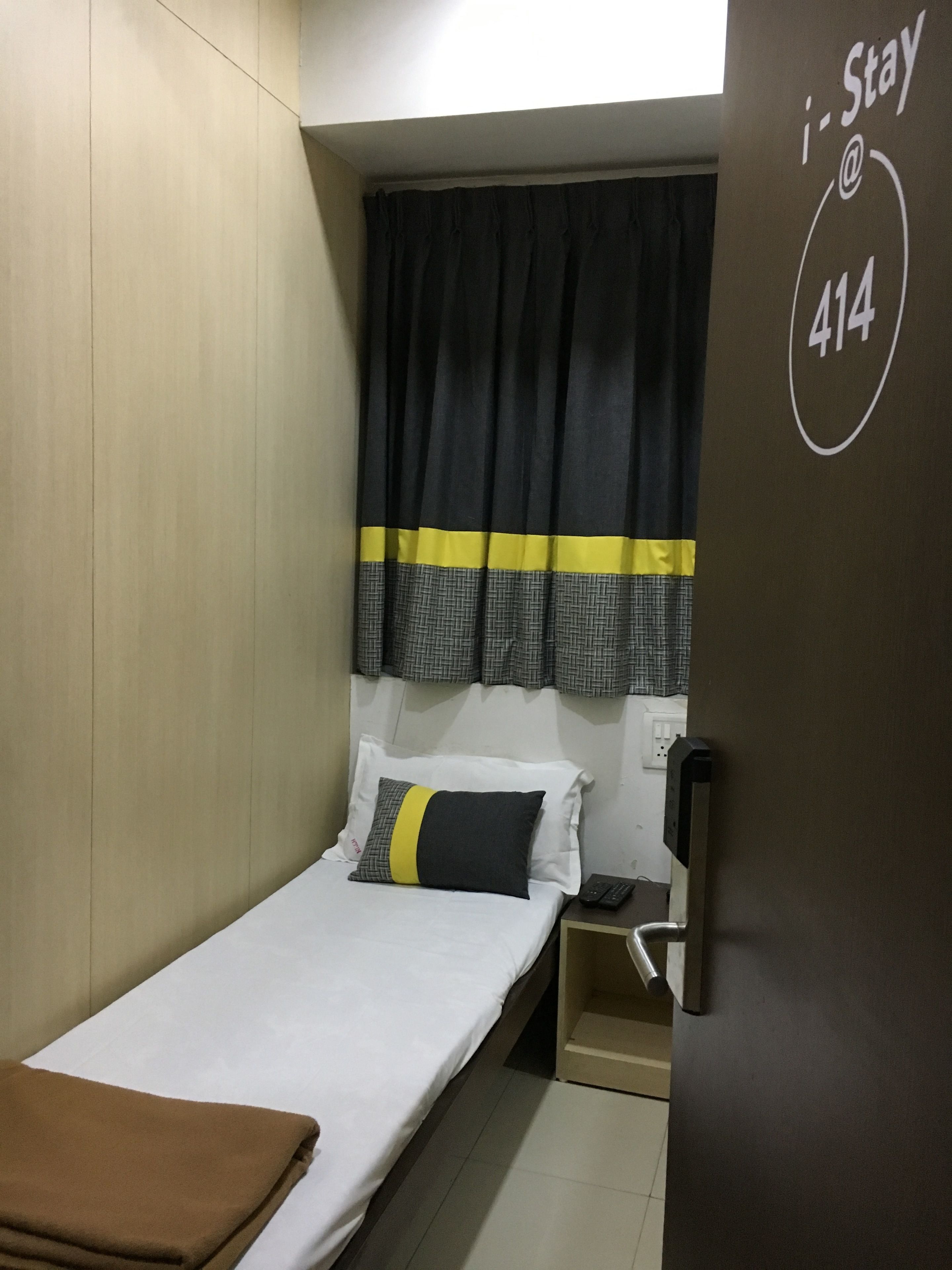 A small, modern hotel room with a single bed, a nightstand, a wall-mounted phone, and a window with black and yellow curtains.