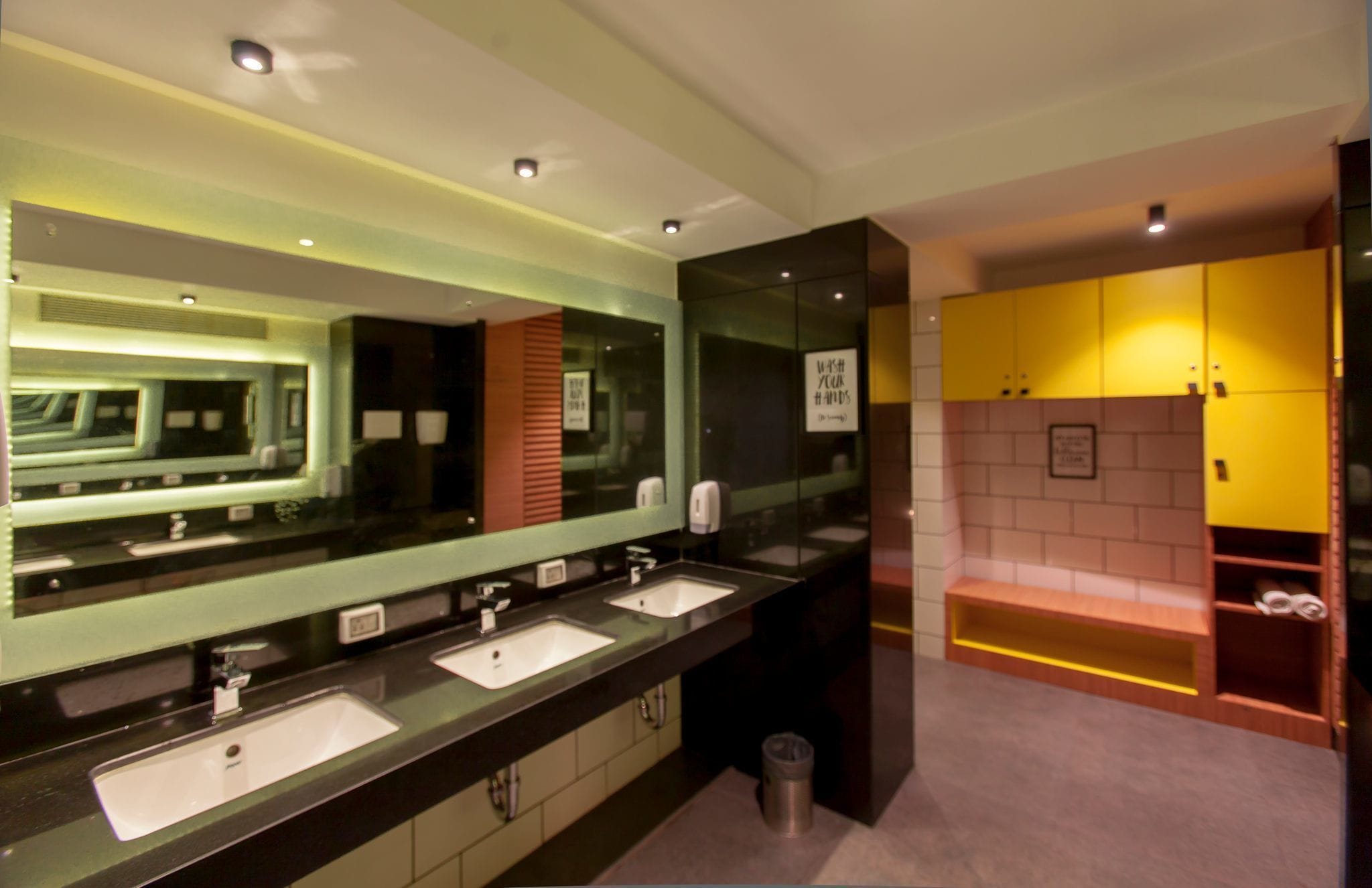 Une salle de bain moderne avec trois lavabos, un grand miroir et un espace douche doté de casiers jaunes.