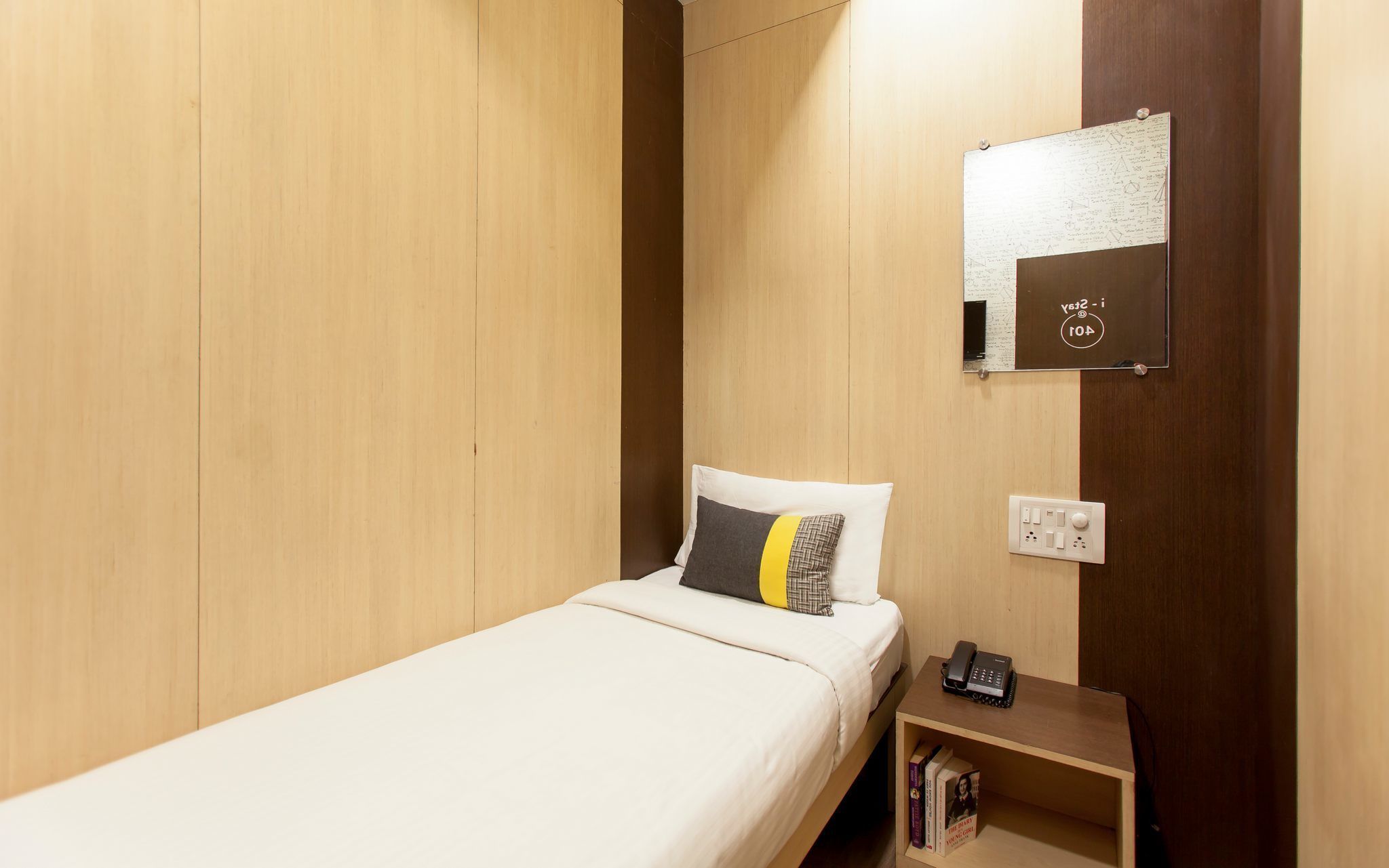 Uma cama de solteiro com colchão e almofadas brancas, uma mesa de cabeceira com telefone e livros, e um espelho fixado na parede com moldura decorativa.