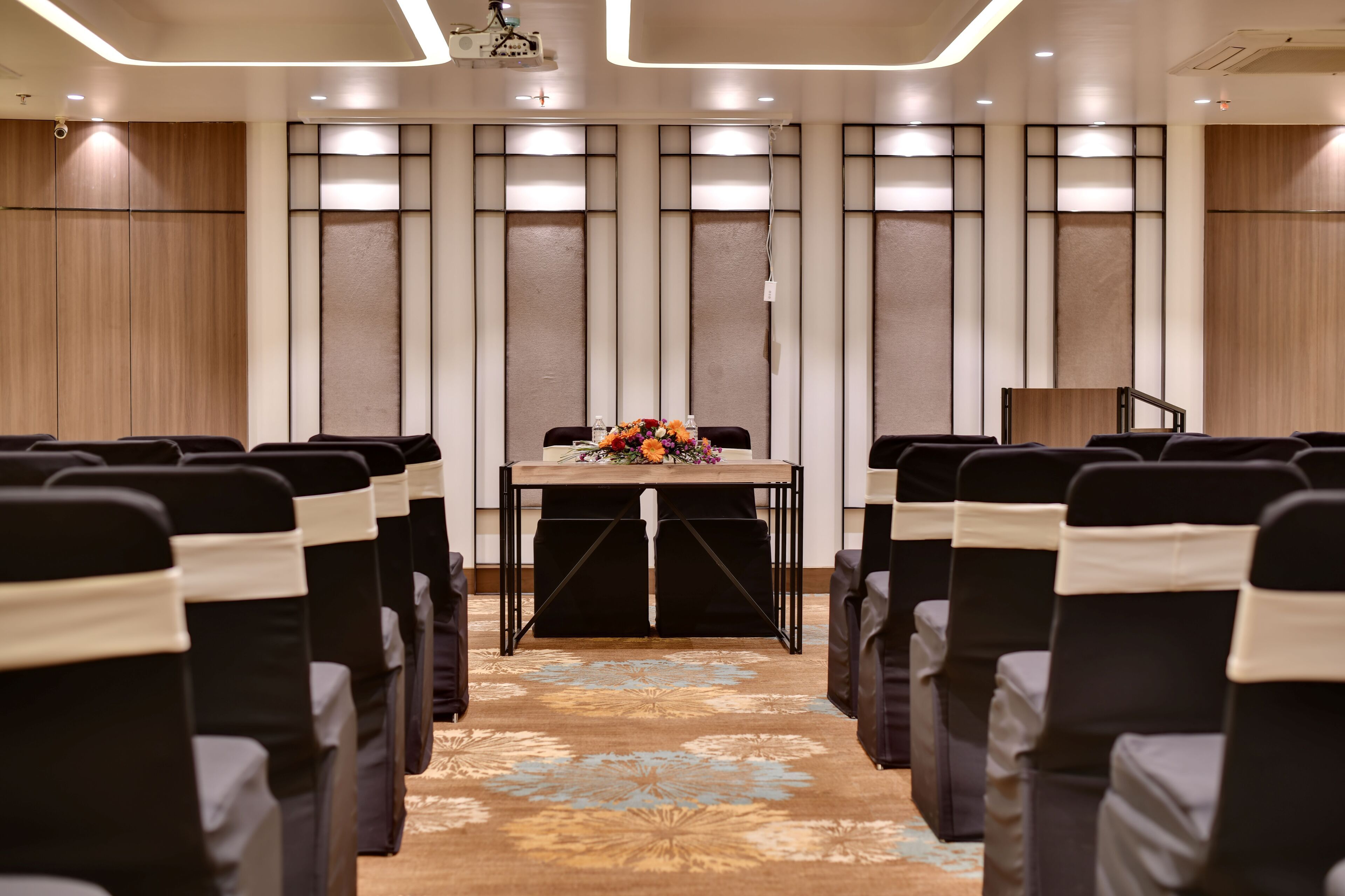 Une salle de conférence avec des rangées de chaises, une table agrémentée d’un arrangement floral et un écran de projection.