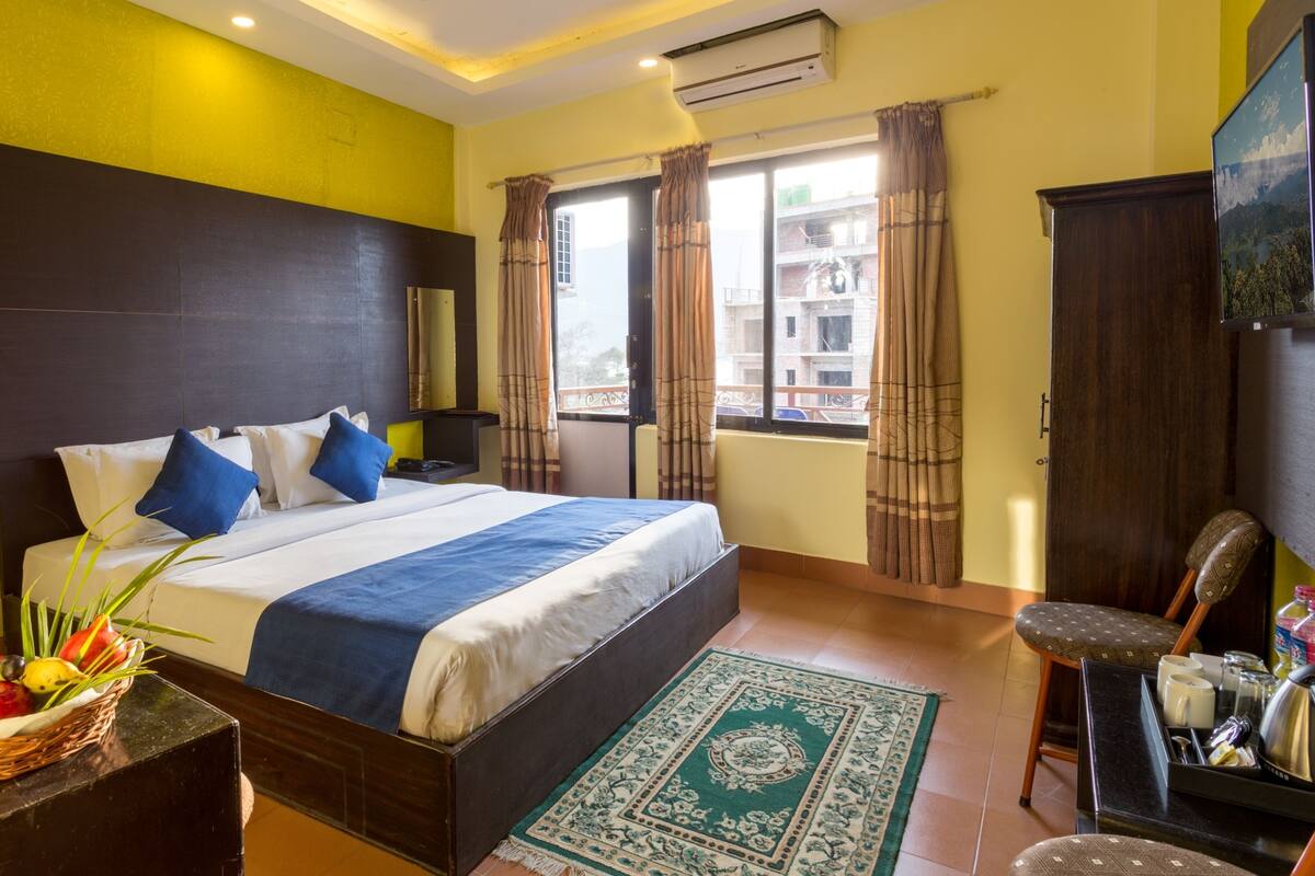 A hotel room with a large bed, a desk with a basket of fruit, a chair, and a view of buildings through the window.