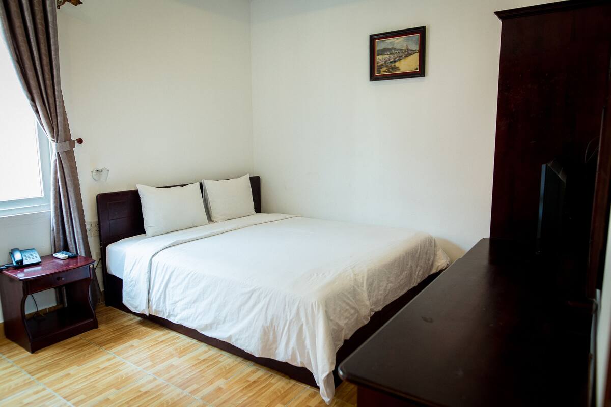 A bedroom with a bed, a nightstand with a phone, a wooden dresser, and a painting on the wall.