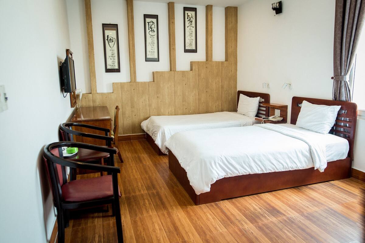 A hotel room with two beds, a wooden floor, a desk with a chair, and framed calligraphy on the wall.