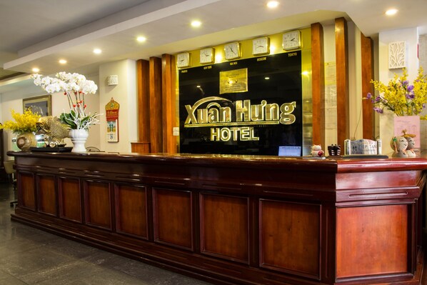 A reception desk with a sign for Xuan Huo Hotel, featuring a clock, a framed certificate, and a vase with flowers.