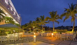 An outdoor dining area with tables and chairs, palm trees, and a building with classical architecture in the background. - Le Beau Max Resort (Fanlu)