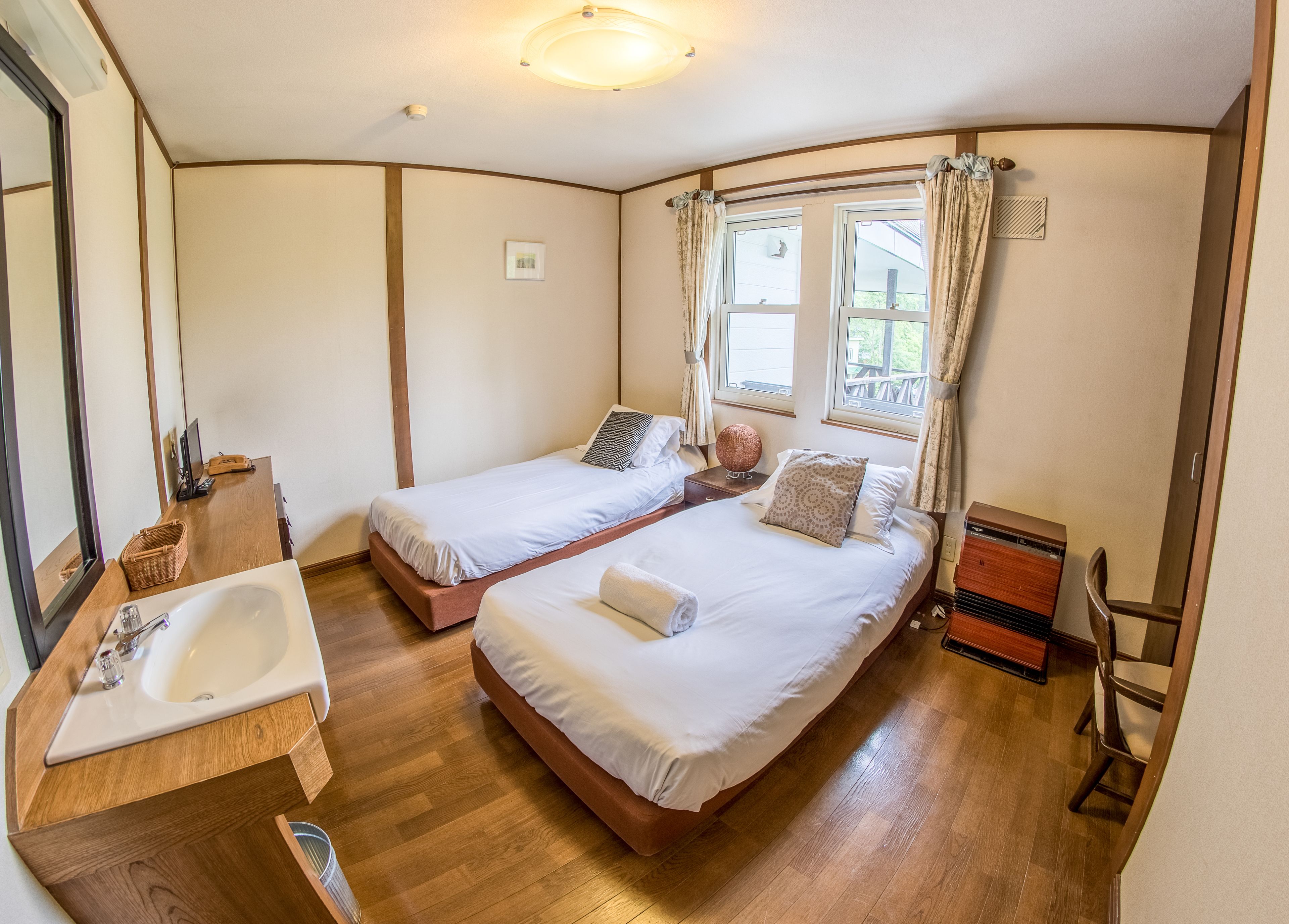 A room with two single beds, a wooden floor, a large window with curtains, a wooden nightstand, a small table, and a bathroom with a sink and mirror.