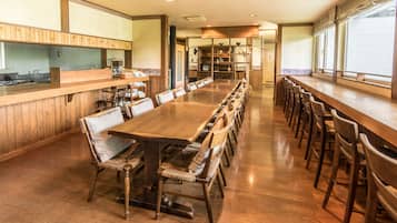 A long wooden dining table with chairs, a kitchen area with wooden cabinets, and large windows.
