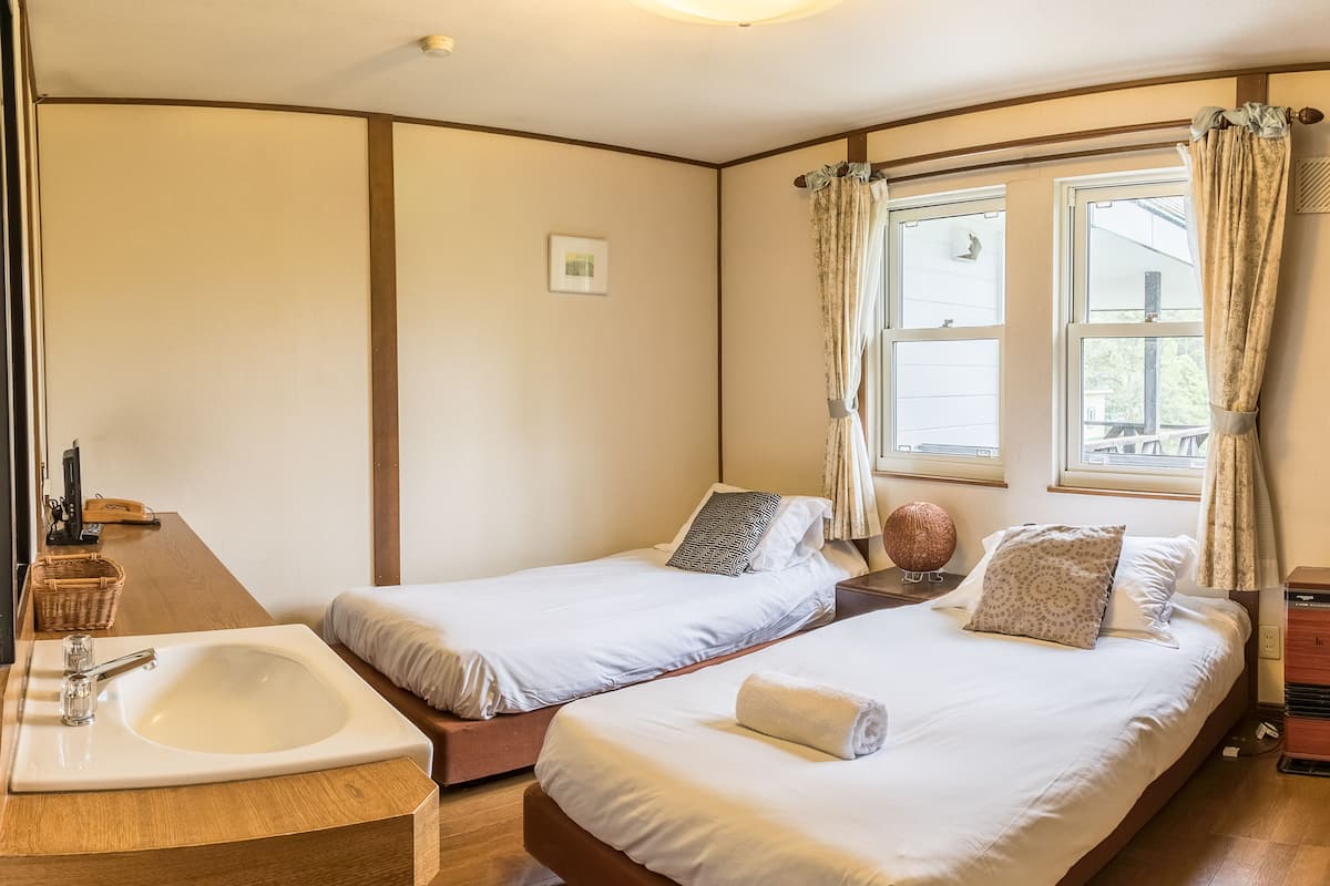 A hotel room with two single beds, a wooden floor, a bathroom with a sink, and a window with curtains.