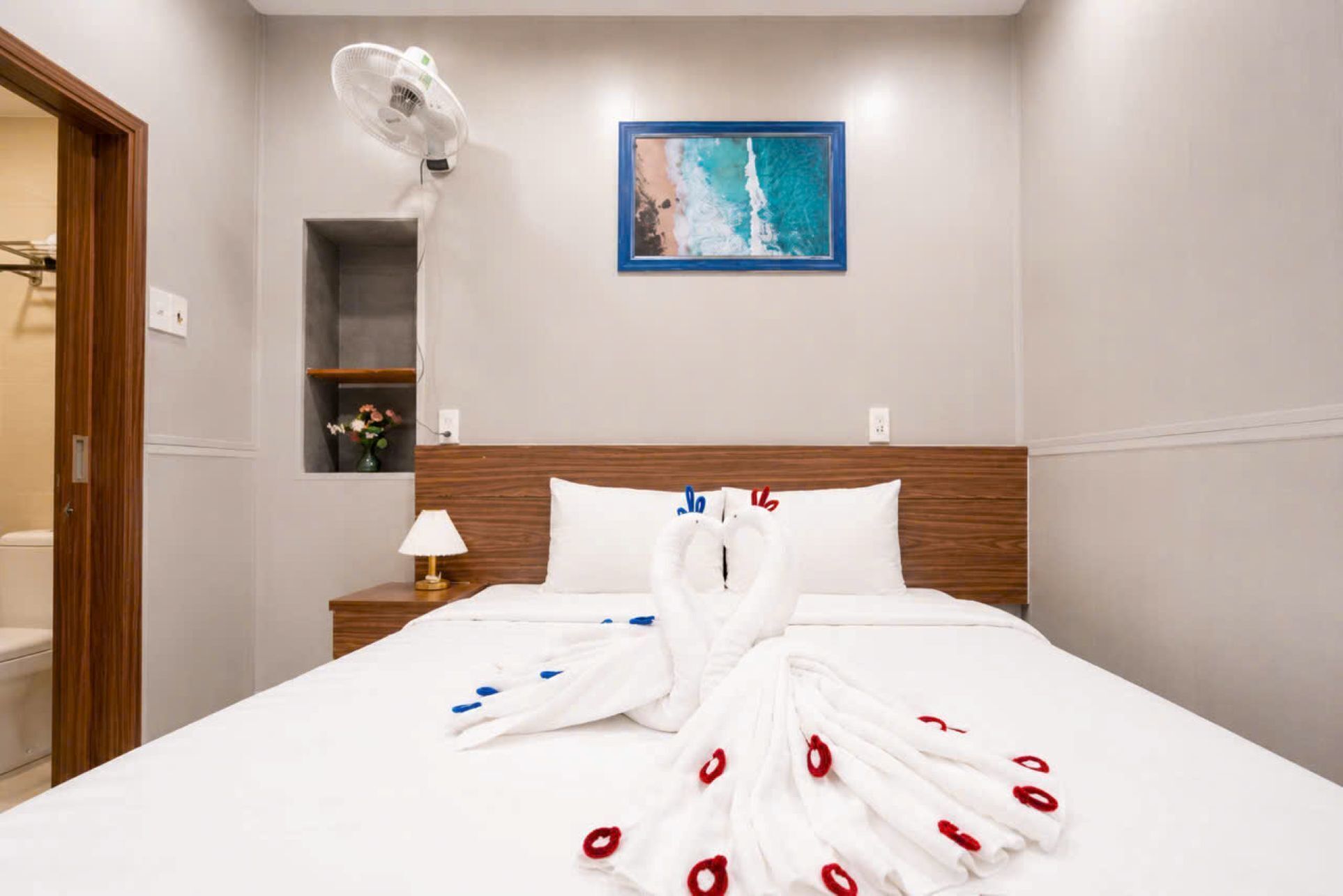 A hotel room with a bed, a wall-mounted fan, a painting, and a small shelf with flowers.