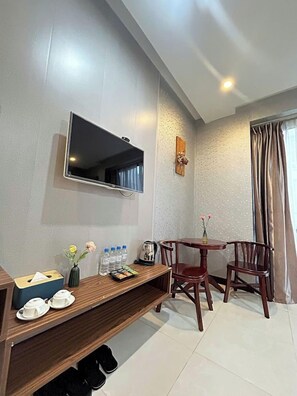 A hotel room with a flat-screen TV mounted on the wall, a wooden console table with a vase, bottles, and a tray of snacks, two wooden chairs, and a pair of slippers on the floor. - BeSea Hotel Da Nang (Da Nang)