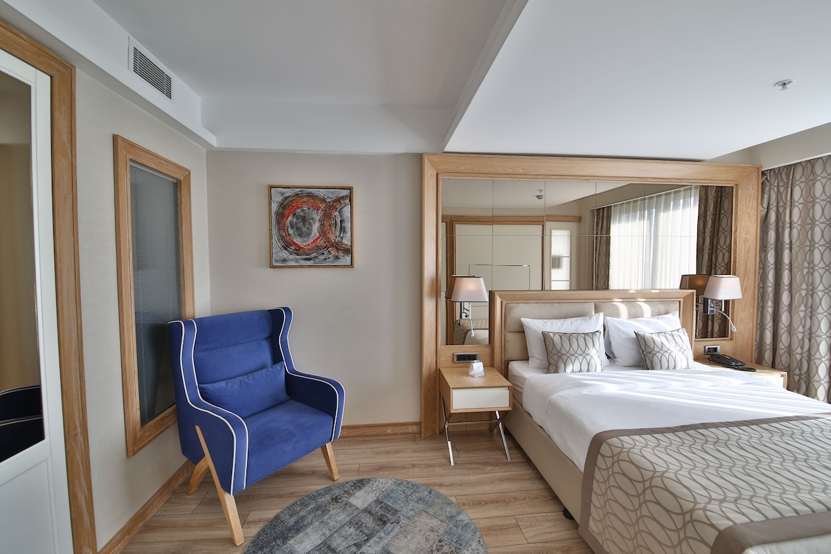 A hotel room with a large bed, a blue armchair, a small table with a lamp, and a large mirror.