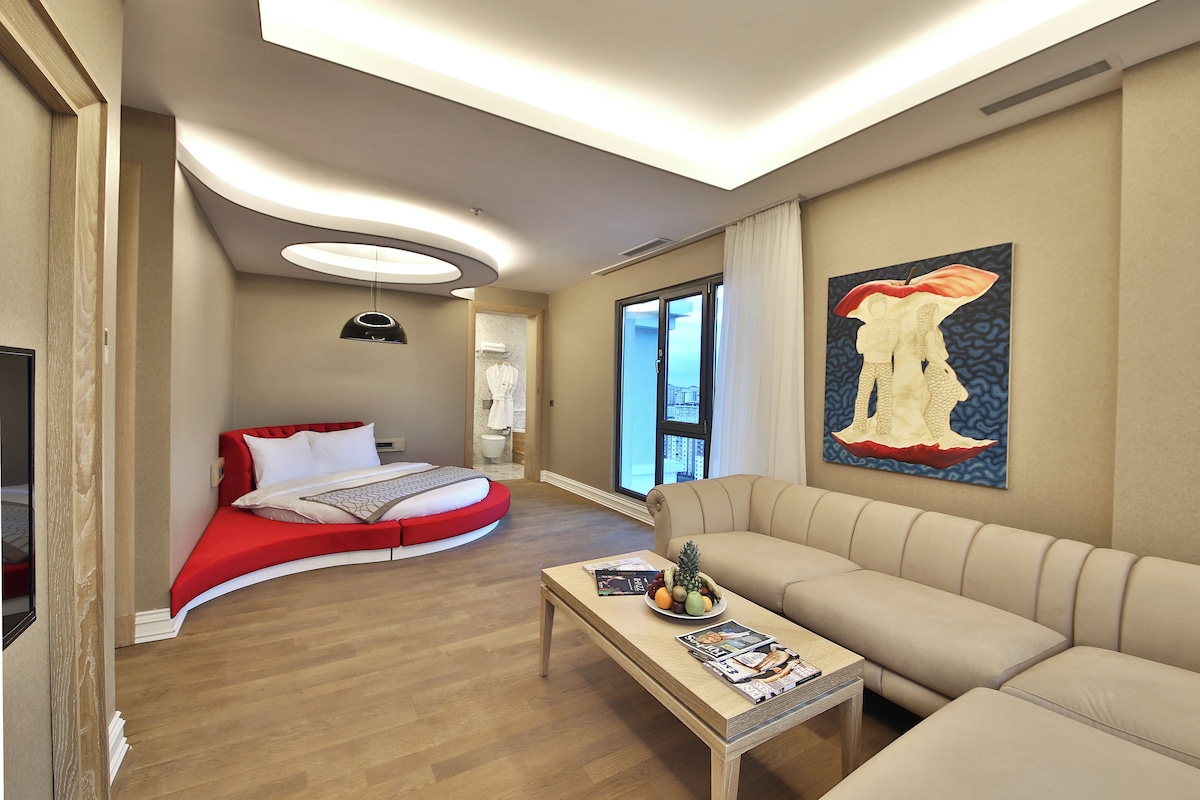A modern hotel room with a curved bed, a sofa, a coffee table with magazines, and a large window with a view.