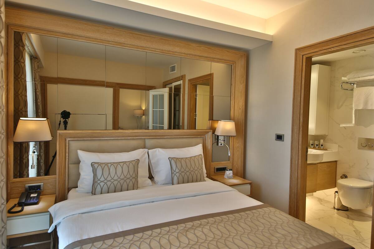 A hotel room with a large bed, bedside lamps, a nightstand with a phone, a mirror above the bed, and a view into the bathroom.
